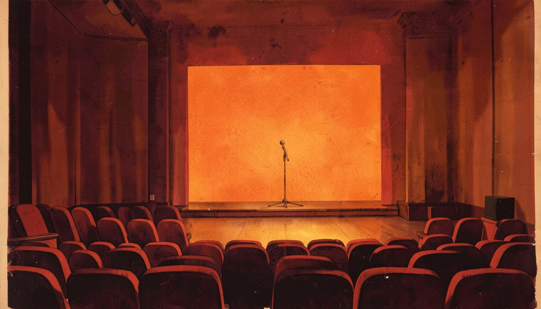 A warm, dimly lit theatre interior with deep amber and burgundy tones, a single microphone on a bare stage, and rows of empty red seats fading into shadow.